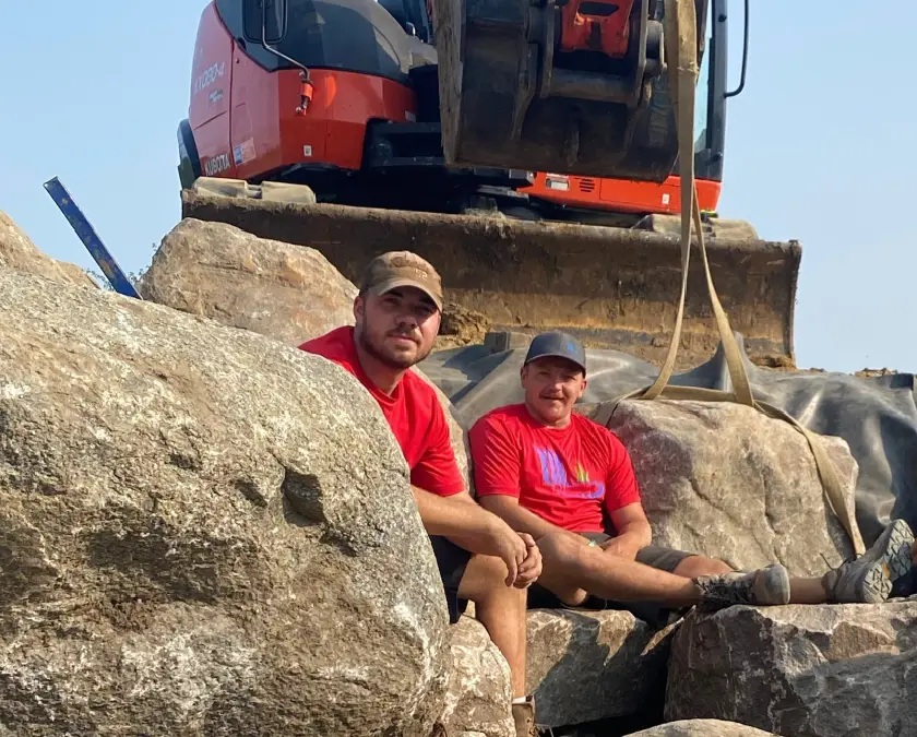 Two Just Add Water crew members resting on boulders in front of an excavator