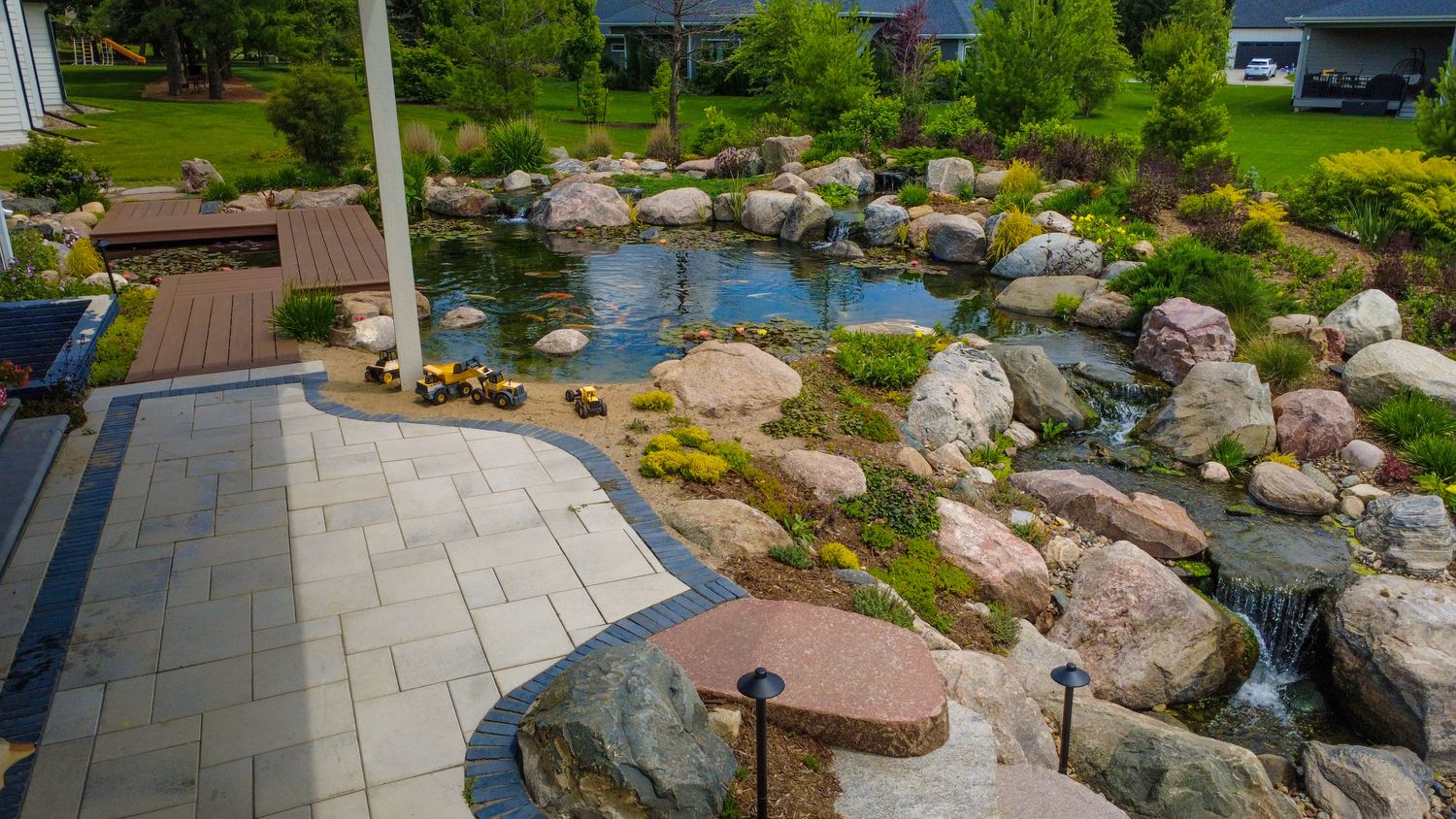 Backyard pond and stream beside a paver patio with a pergola and low lights