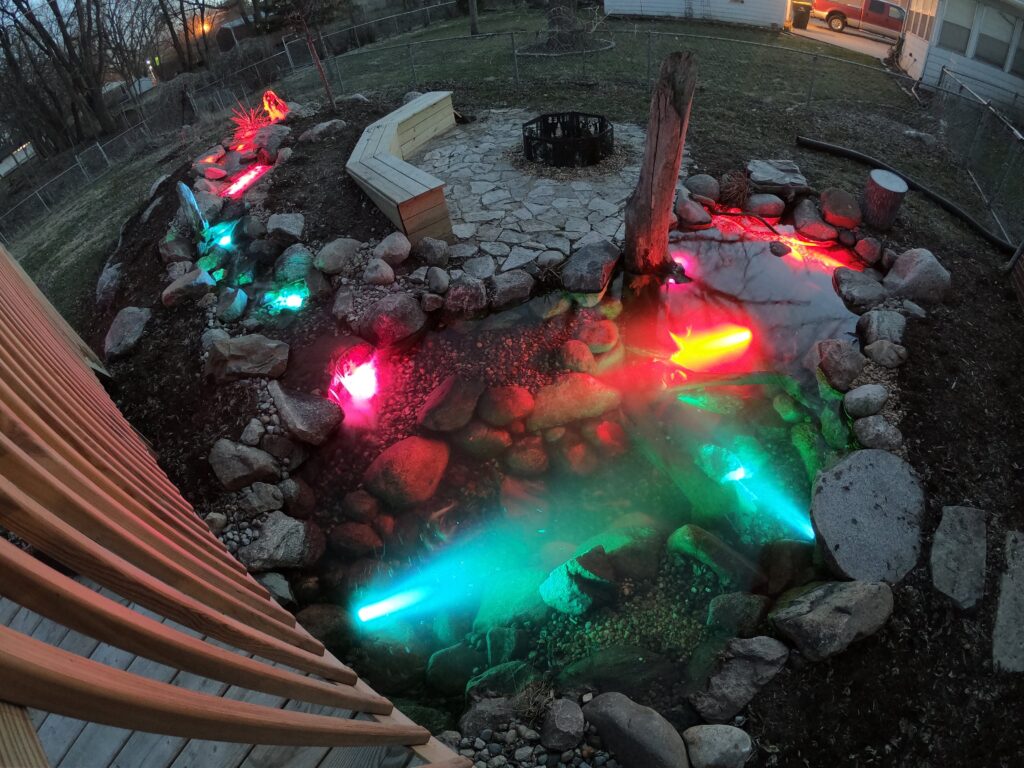 Pond lit with red and teal underwater lights next to a fire pit at night