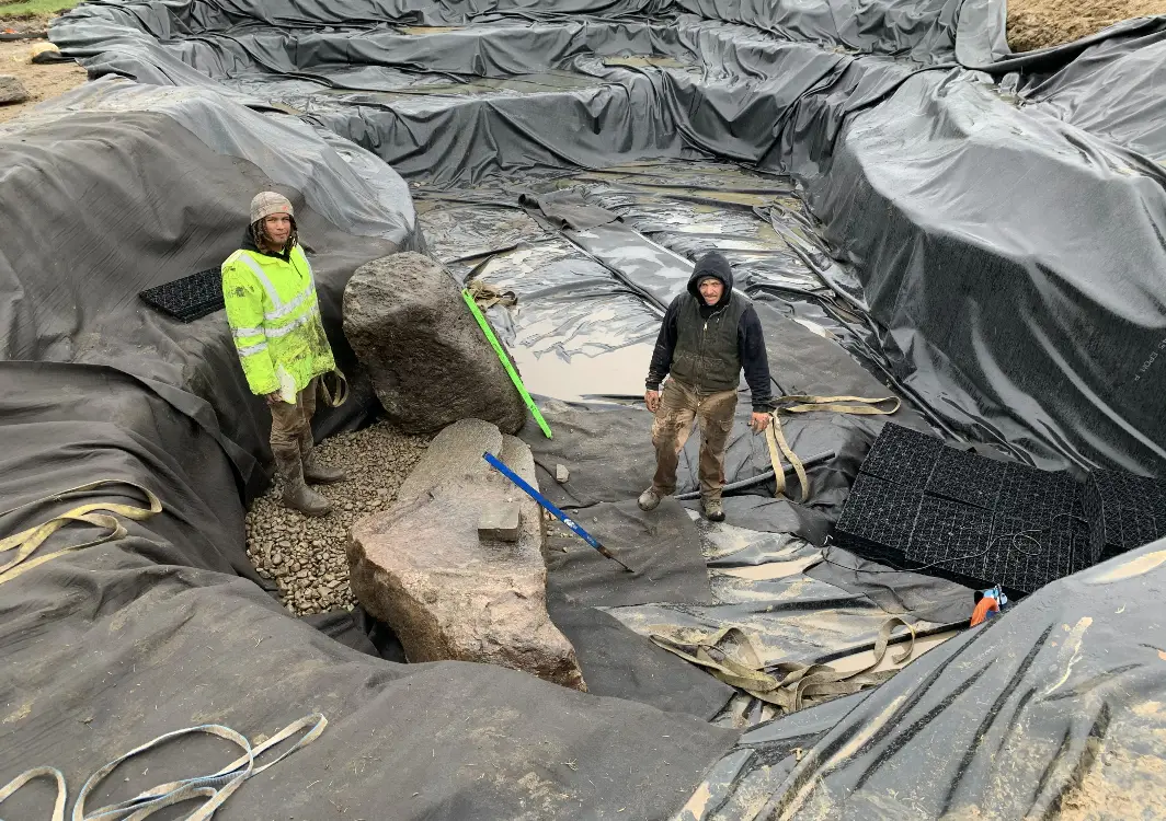 Two Just Add Water crew members standing on underlayment inside a large pond build