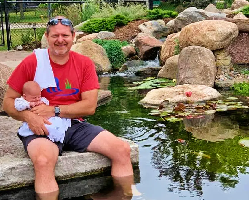 Just Add Water owner Chris holding a baby while sitting at the edge of a pond