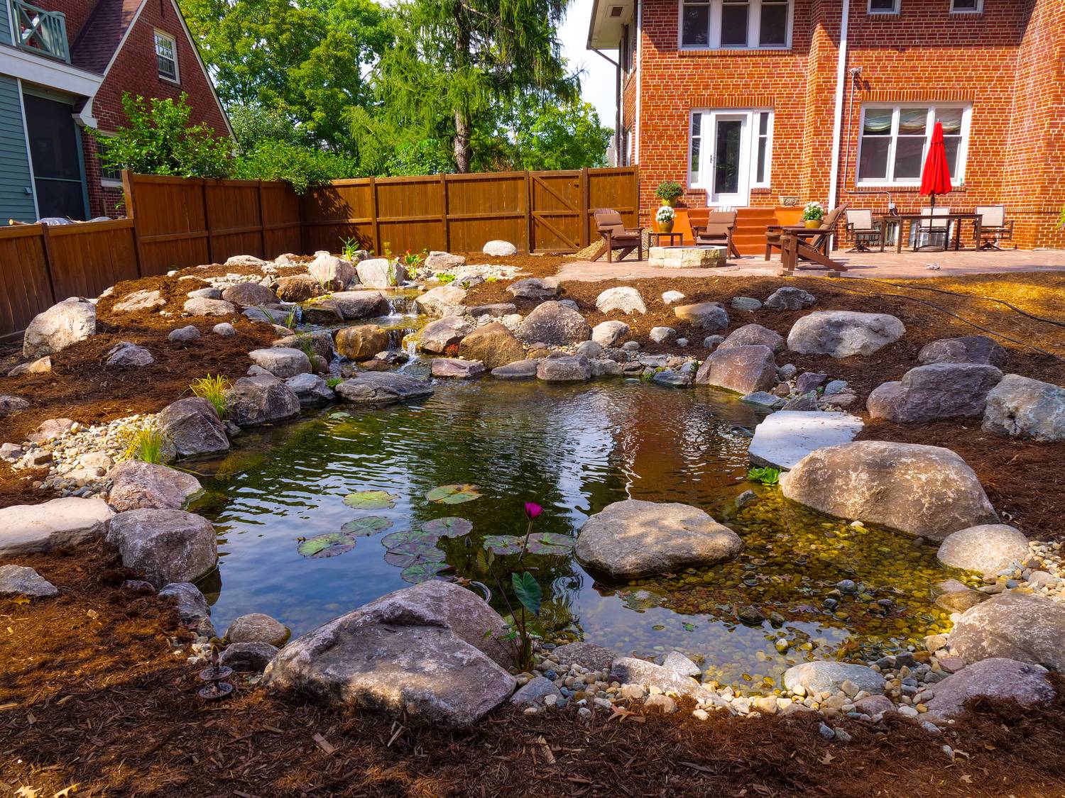 Newly built boulder pond in front of a brick house with patio seating