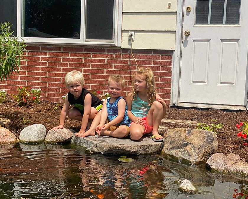 Three children sitting on a flat rock at the edge of a backyard pond