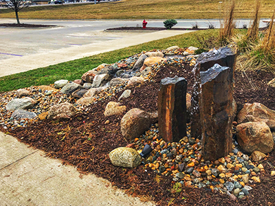 Three basalt column fountains set in river rock and mulch by a sidewalk
